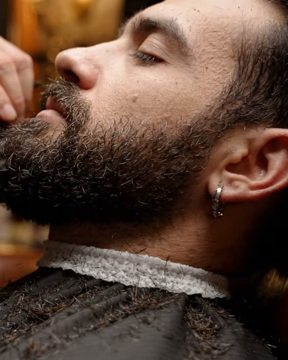 Cliente de olhos fechados, recostado na poltrona, enquanto o barbeiro apara a barba com tesoura e pente