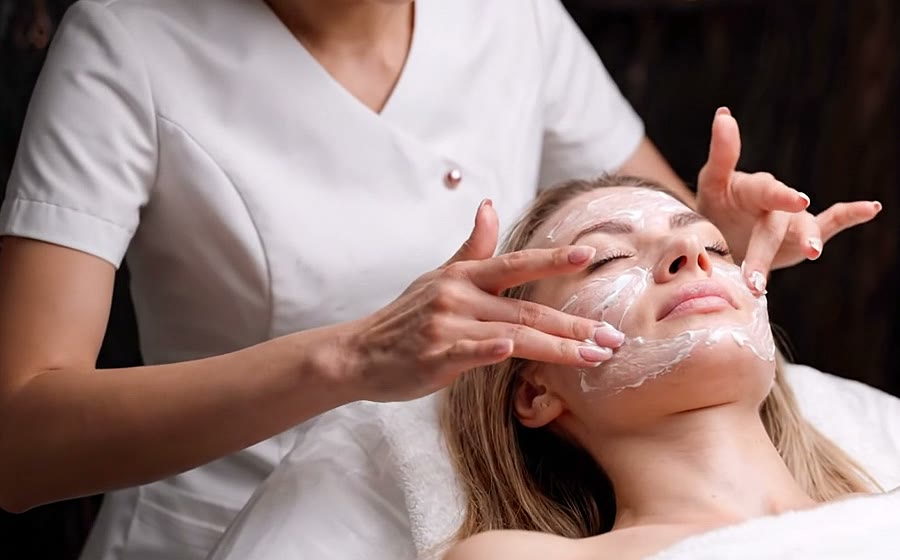 Esteticista de jaleco branco aplica creme no rosto de uma cliente deitada na maca