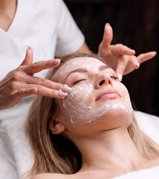 Esteticista aplica creme no rosto de uma cliente deitada na maca, com movimentos suaves das mãos