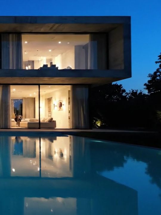 Fachada de vidro iluminada ao lado de árvores, com a piscina em primeiro plano