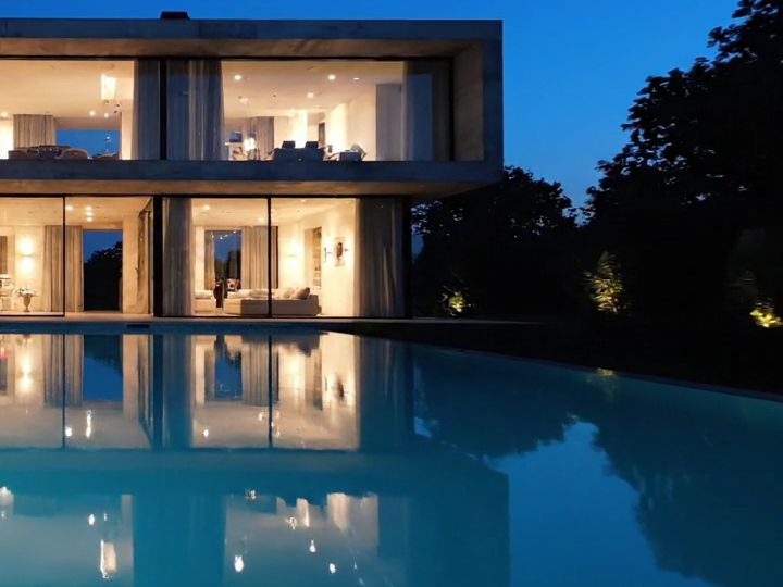 Sobrado iluminado à noite, com jardim arborizado e piscina refletindo as janelas