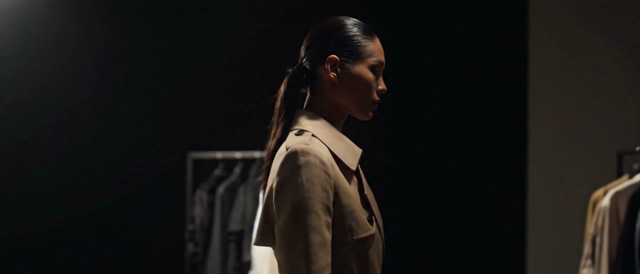 Modelo de trench no ateliê escuro, com uma arara de roupas ao fundo e uma parede clara à direita