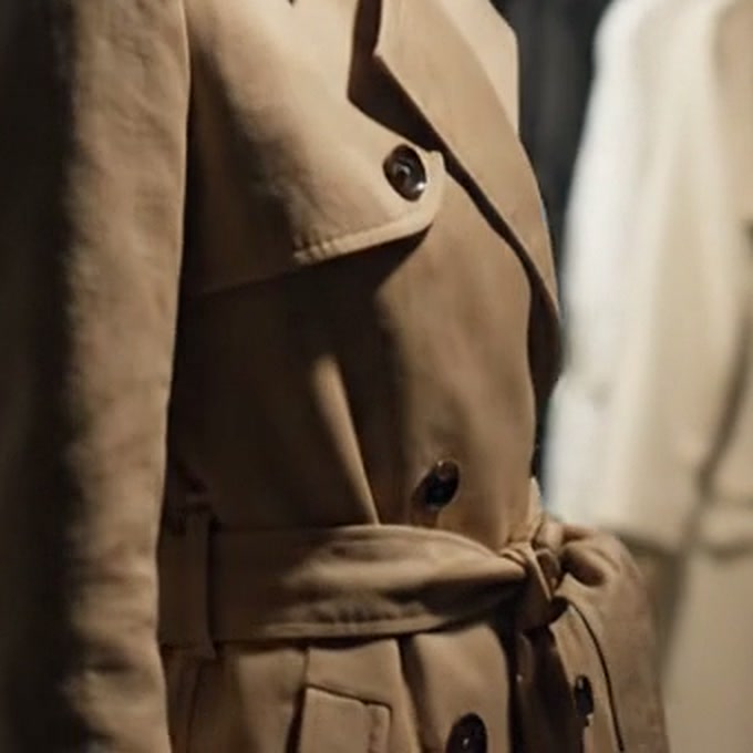 Detalhe do cinto amarrado e dos botões escuros do trench, com casacos claros desfocados ao fundo