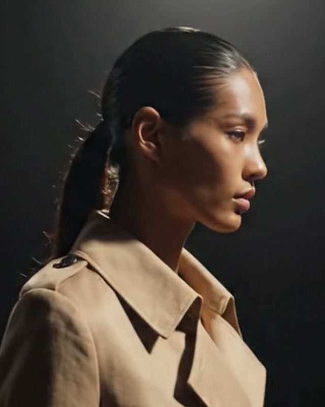 Rosto de perfil da modelo, de cabelo preso, com a gola do trench areia levantada