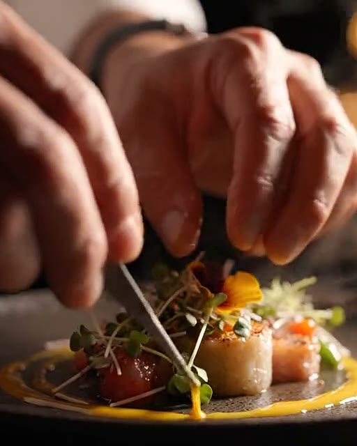 Mãos do chef finalizando um prato com pinça, à luz de velas